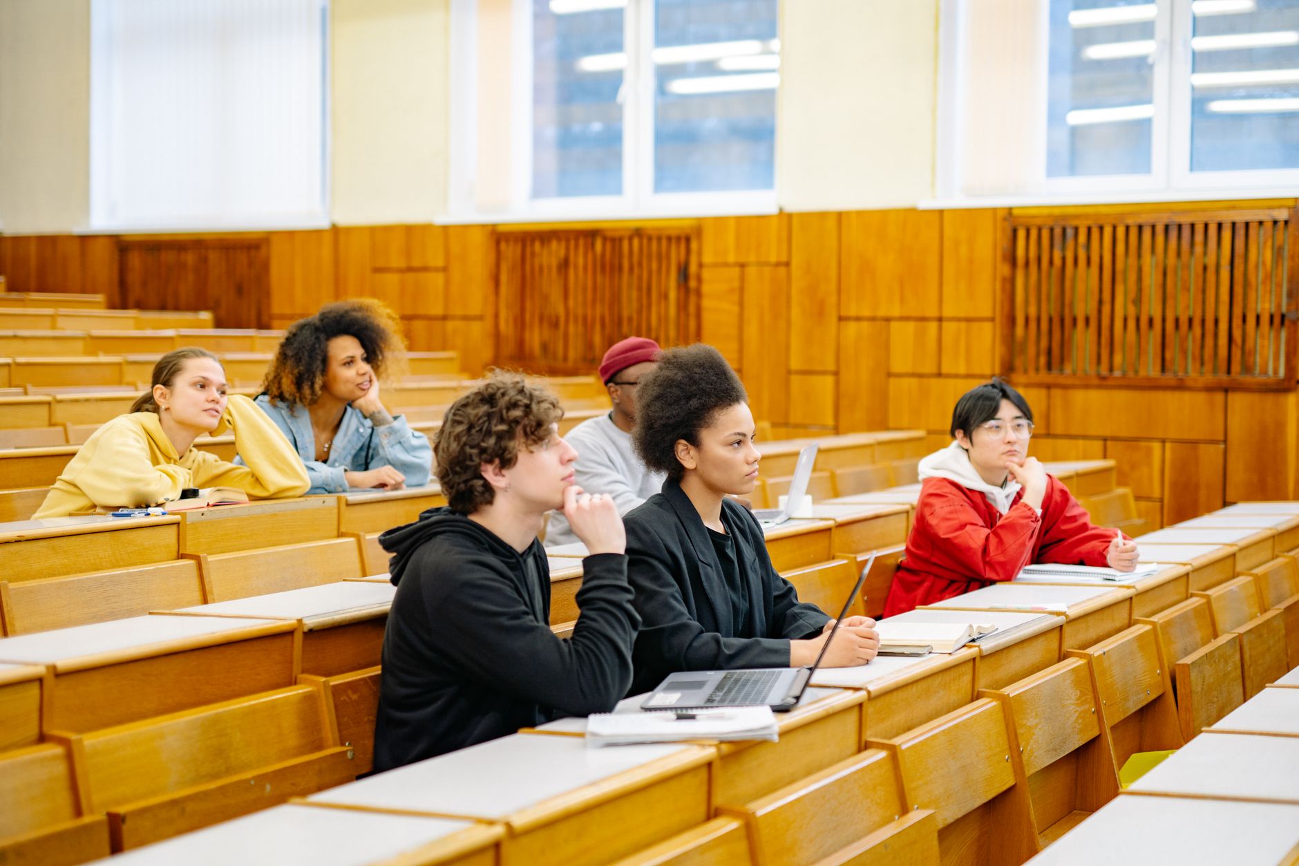 Cinq étudiants assis dans un amphithéâtre en bois, écoutant avec attention, ordinateurs portables et cahiers posés sur les pupitres.