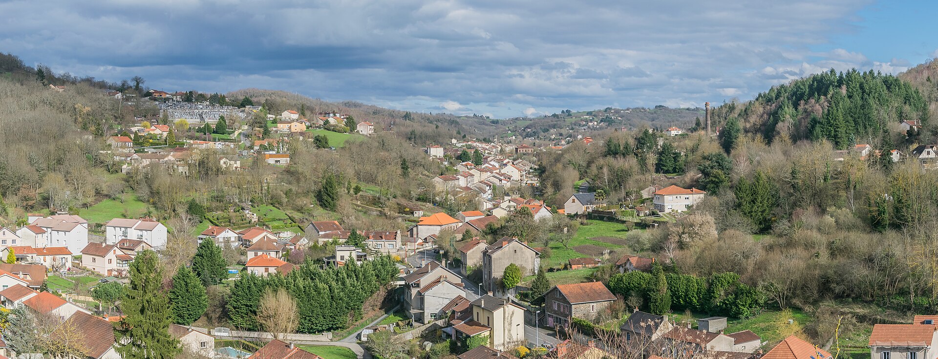 Panorama de la vallée d'Aubin, avec ses maisons aux toits rouges entre les collines boisées sous un ciel nuageux