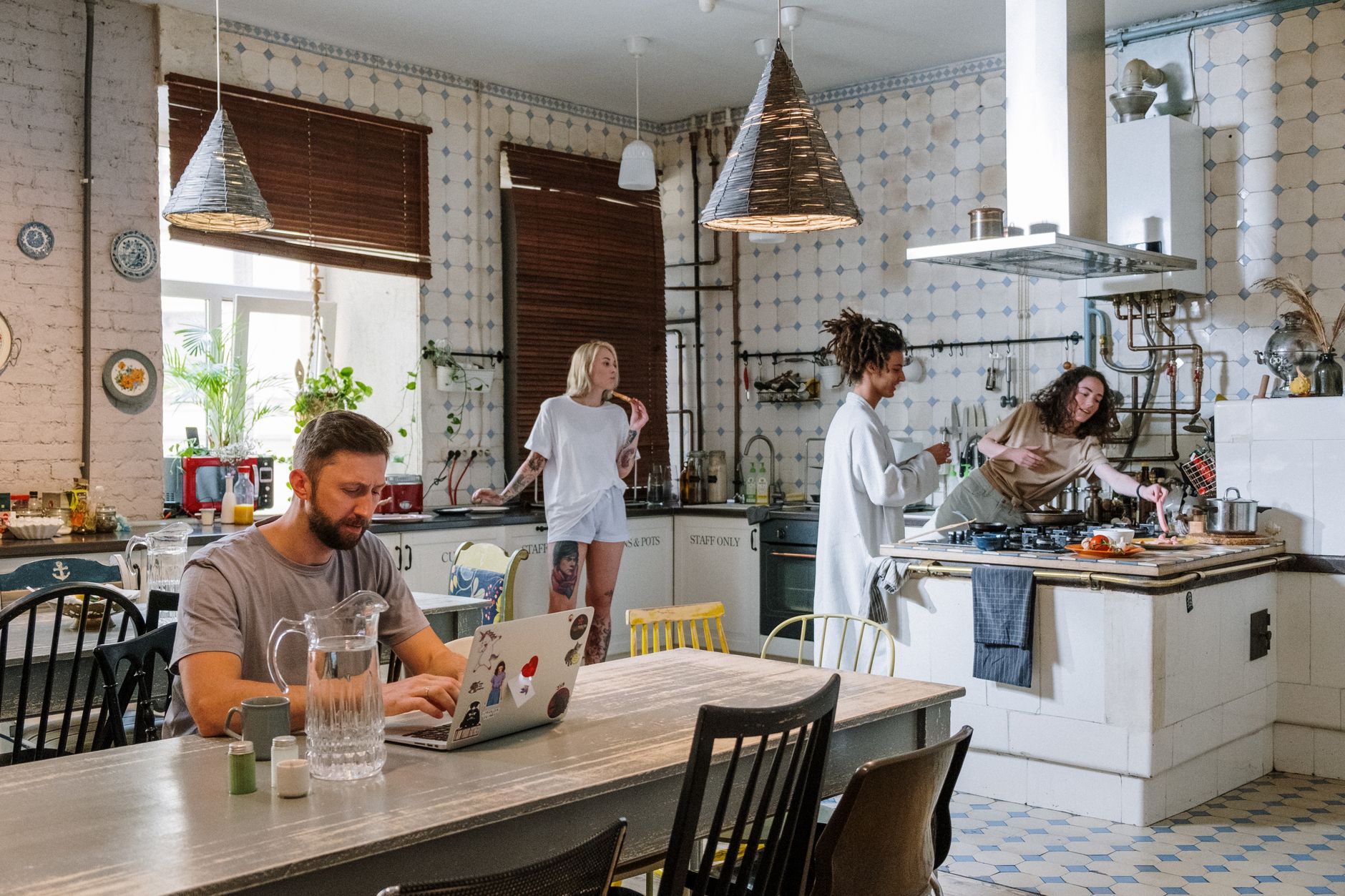 Un homme travaille sur un ordinateur portable à une table de cuisine, pendant que trois autres personnes se tiennent ou cuisinent au fond de la pièce.